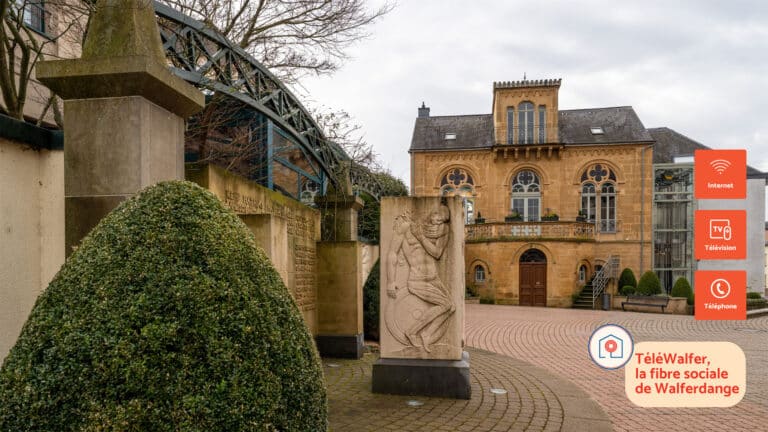 View of Maison Dufaing in Walferdange, a historic 19th-century building with arched windows and ornate stonework. In the foreground, a sculpted stone war memorial and manicured bushes are visible. On the right, modern TéléWalfer icons indicate services such as internet, TV, and telephone, with the slogan “TéléWalfer, la fibre sociale de Walferdange