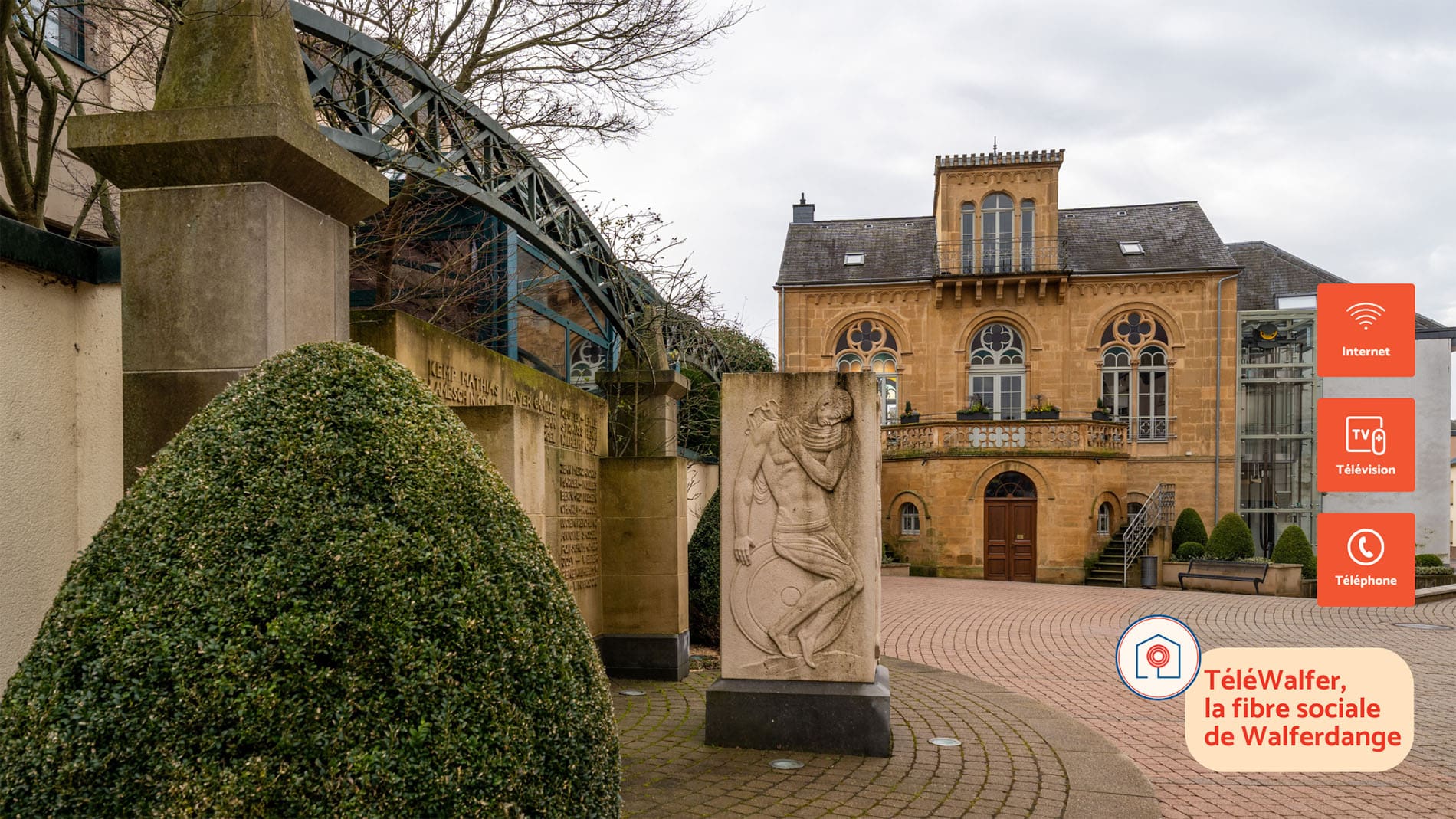 View of Maison Dufaing in Walferdange, a historic 19th-century building with arched windows and ornate stonework. In the foreground, a sculpted stone war memorial and manicured bushes are visible. On the right, modern TéléWalfer icons indicate services such as internet, TV, and telephone, with the slogan “TéléWalfer, la fibre sociale de Walferdange
