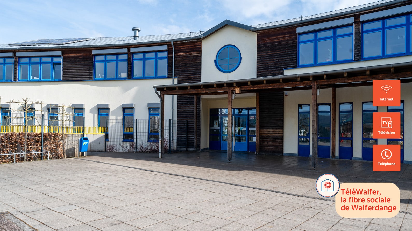 Front view of a modern primary school building in Walferdange with blue-framed windows and wooden accents. The entrance is sheltered by a wooden canopy. On the right, TéléWalfer icons highlight internet, television, and phone services, along with the slogan “TéléWalfer, la fibre sociale de Walferdange.